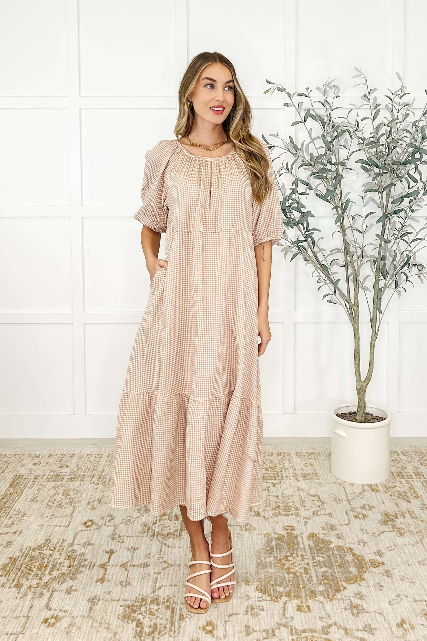 A woman standing in an indoor setting wearing a beige, short-sleeved, tiered dress with a round neckline.