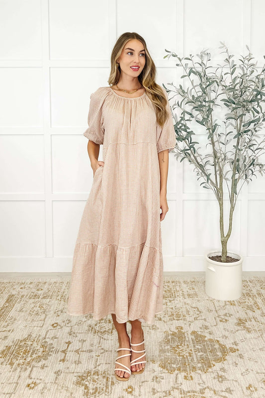 A woman standing in an indoor setting wearing a beige, short-sleeved, tiered dress with a round neckline.