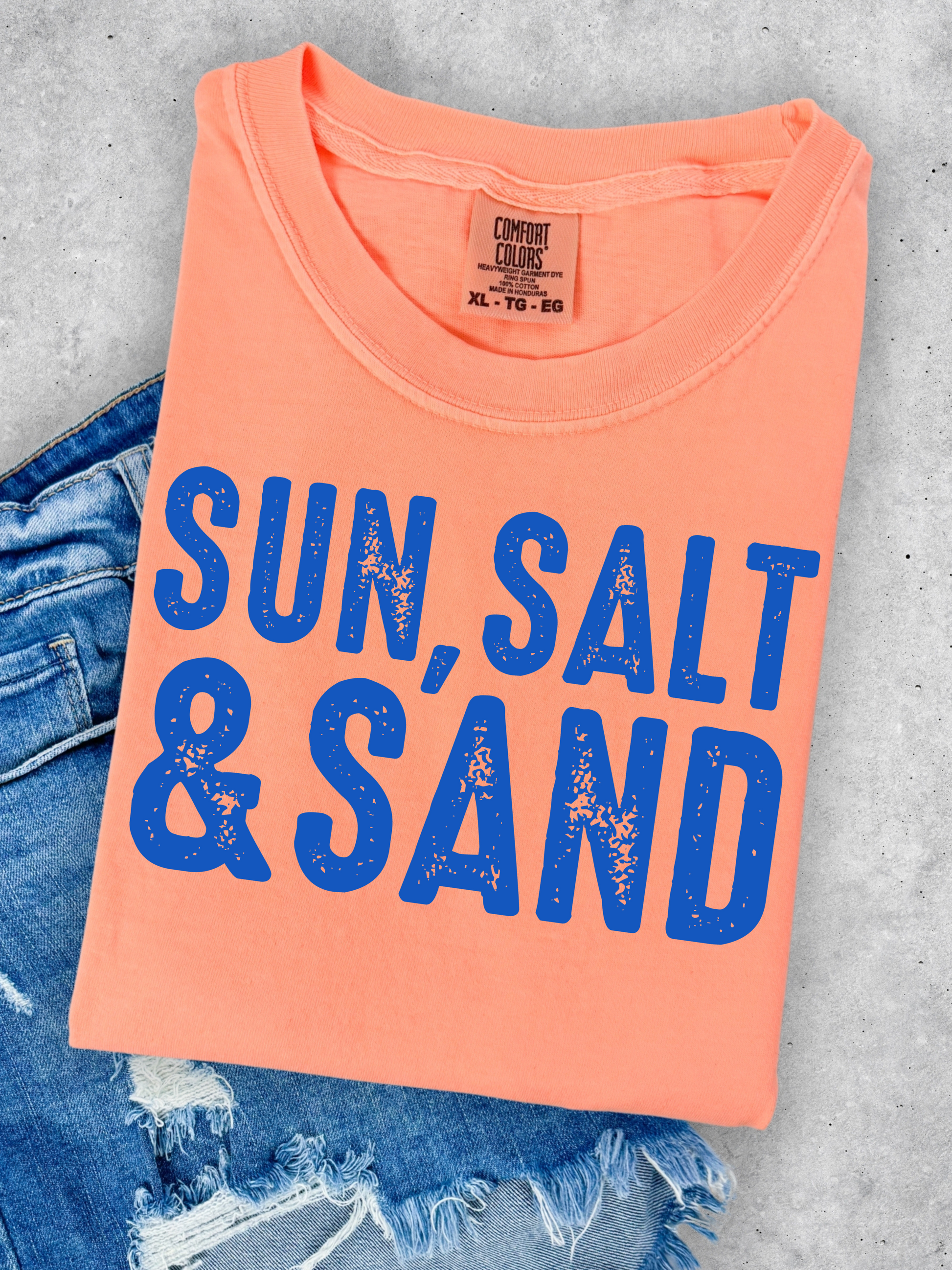 Folded peach t-shirt with 'Sun, Salt & Sand' text on a gray surface with denim jeans.