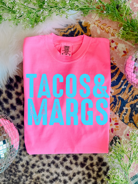 Pink t-shirt with 'TACOS & MARGS' text on a patterned surface with decorative elements.