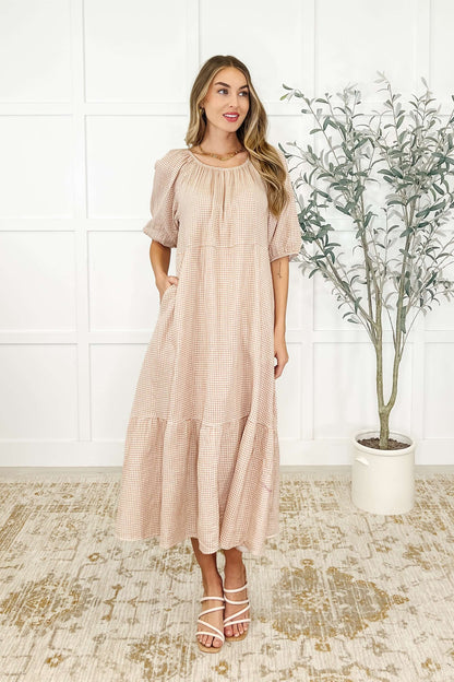 A woman standing in an indoor setting wearing a beige, short-sleeved, tiered dress with a round neckline.